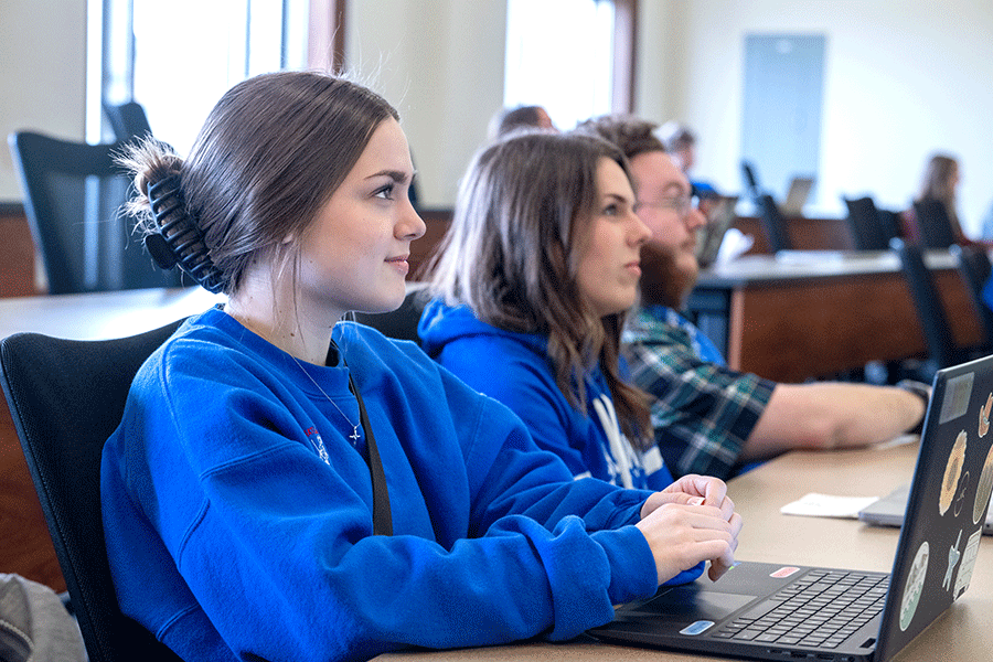 Three students – two women with brown hair in blue sweatshirts, and a man with facial hair and a checkered shirt – sit in front of laptops as they look toward the front of a classroom. The woman nearest the camera is smiling softly.